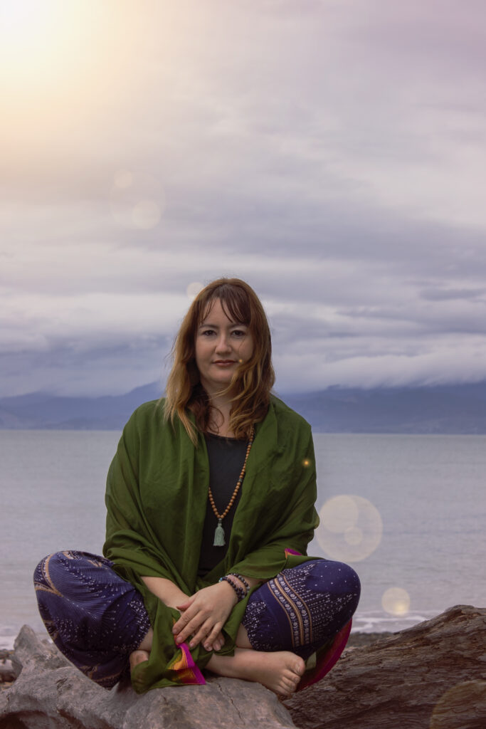 Debbie Sumner seated in meditation in a natural seaside setting at sunset during a yoga practice in Mapua